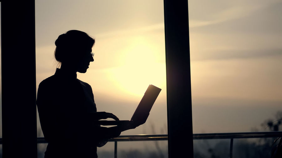 Dark silhouette of business woman, typing something in laptop, holding it in hands