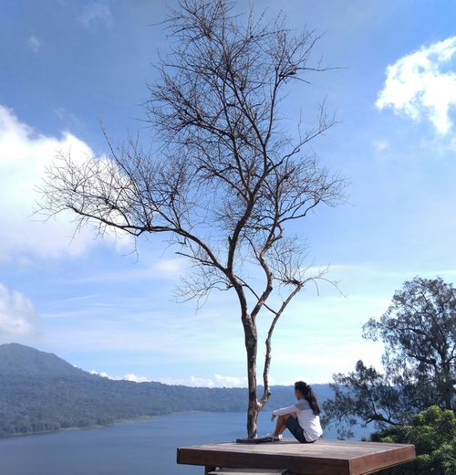 Man sitting on tree by lake against sky