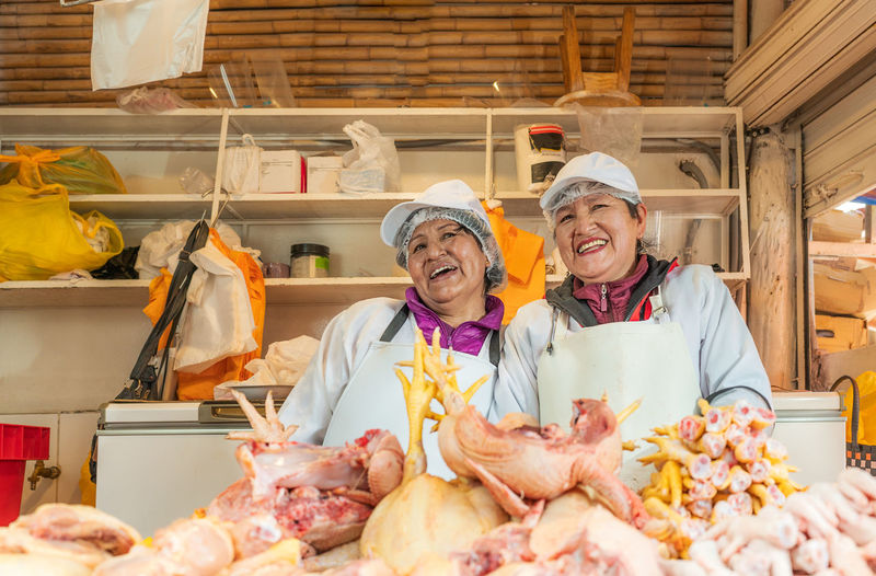 Cheerful peruvian female sellers in aprons looking away while standing behind counter with raw chicken meat during work on itinerant fair
