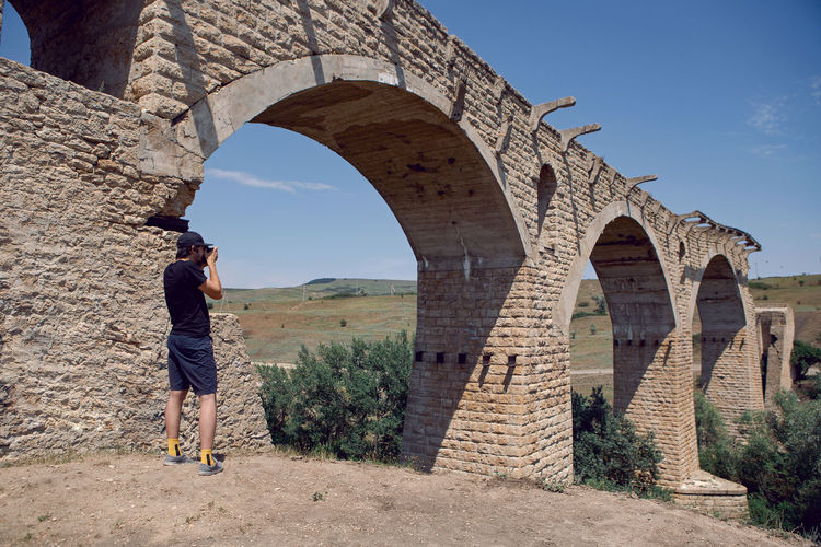 Guy traveler in a cap takes photos of a high stone bridge destroyed