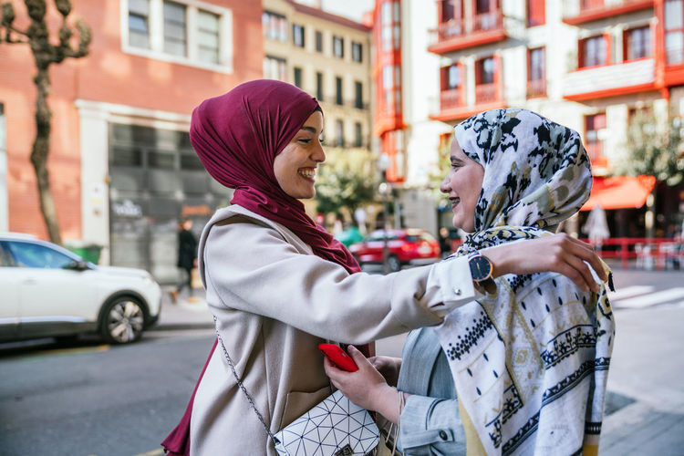 Side view of content muslim female friends in hijabs standing in street and hugging while looking at each other