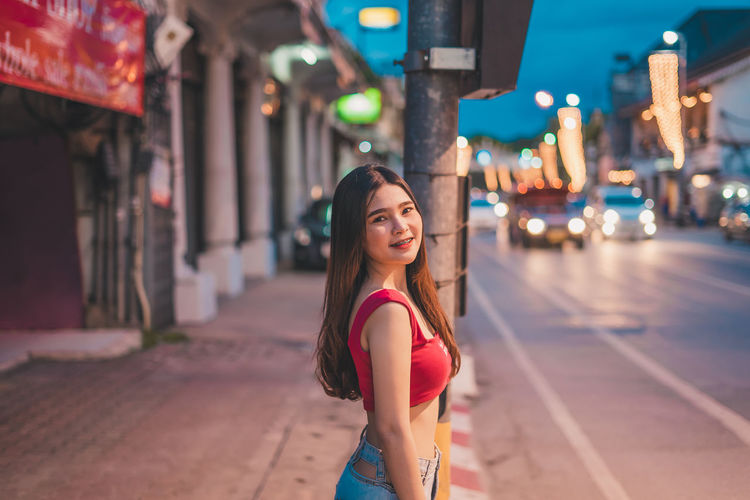 Portrait of smiling young woman standing on street in city at dusk
