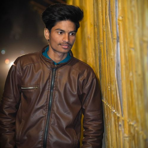 Young man looking away while standing against fence at night