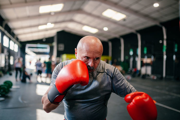 Powerful serious adult mature male boxer in red gloves looking away with challenge while training in boxing gym