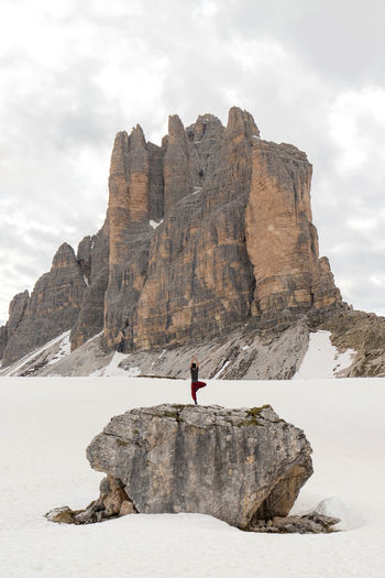 Remote view of traveler standing in tree pose on rock and practicing yoga in snowy dolomites mountains