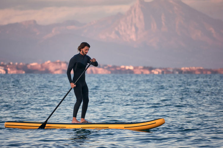 Side view of male surfer in wetsuit rowing on paddle board on sea water on background of mountains at sunset