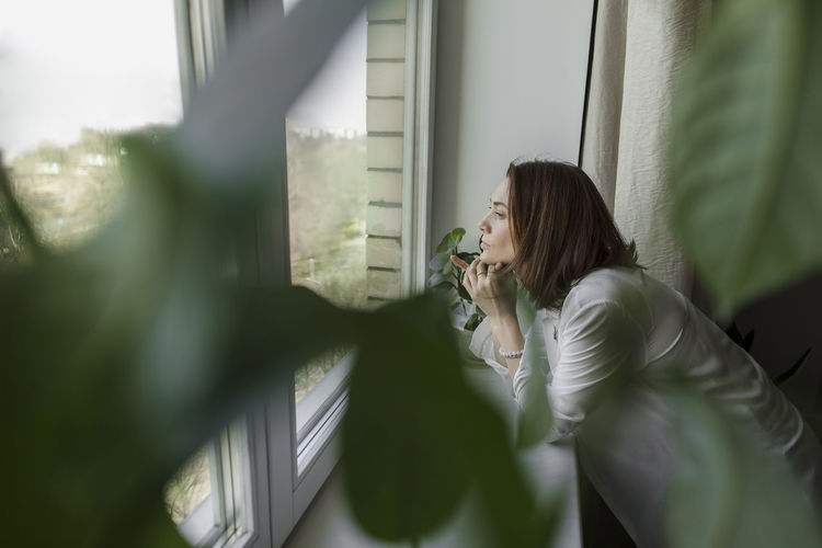 Thoughtful woman with hand on chin looking through window at home
