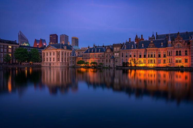 Illuminated buildings by river against blue sky at dusk