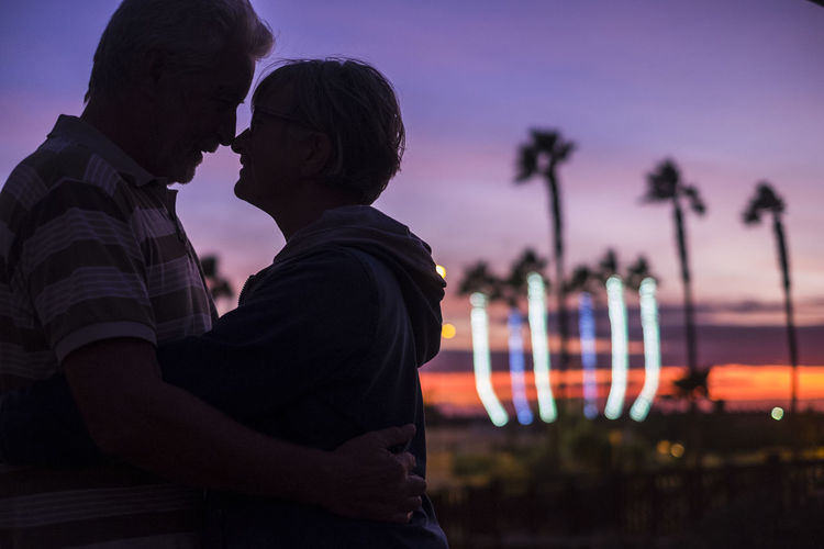 Side view of senior couple embracing against sky during sunset