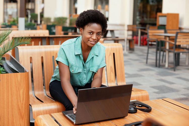 Portrait of smiling young woman using laptop while sitting at sidewalk cafe
