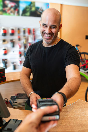 Delighted adult man in black t-shirt smiling and giving payment terminal to crop client while working in bike shop