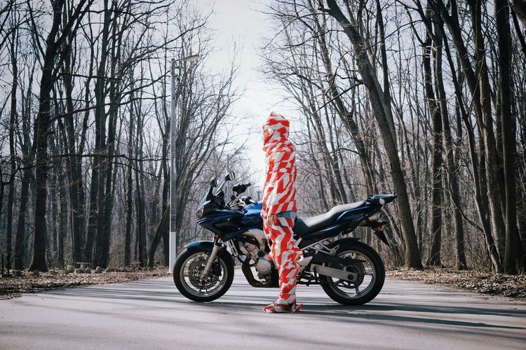 Man covered in red fabric while standing by motorcycle against bare trees