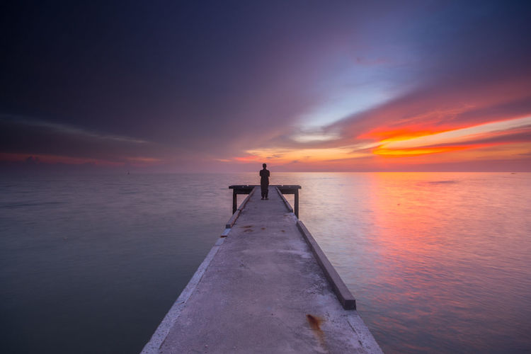 Rear view of man standing on pier over sea against sky during sunset