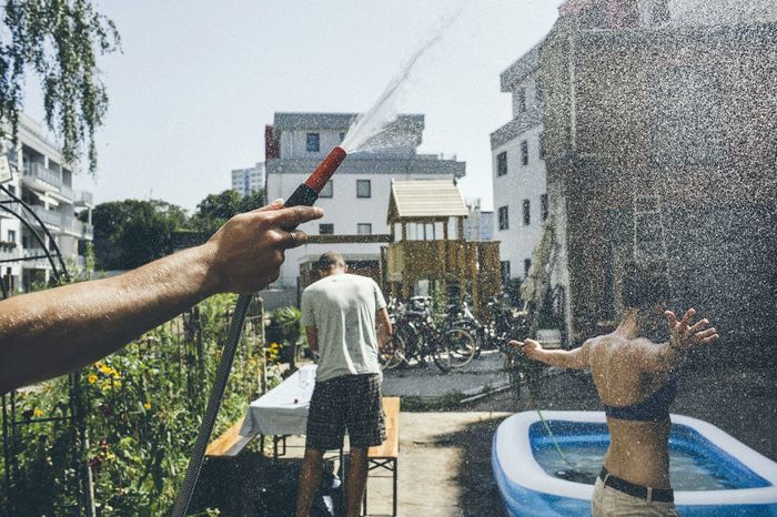 Cropped hand holding pipe with water splashing on woman by buildings against sky