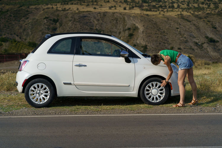 Young woman takes care of car checking rims on wheel against slopes in countryside on sunny day