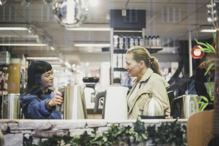 Saleswoman with kettle looking at female customer in store seen through glass window