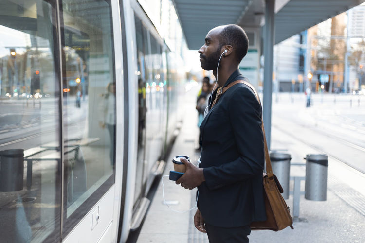 Young businessman with coffee to go standing at tram stop listening music with earphones and smartphone