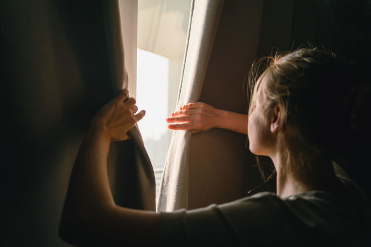 Back view of unrecognizable young female in casual clothes opening blackout curtains and admiring modern city during holidays in hotel