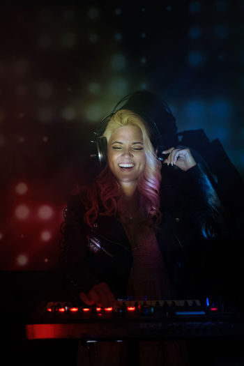 Happy young female dj in headphones smiling and using synthesizer to play music while standing in smoke during party in nightclub
