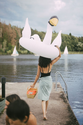 Rear view of teenager carrying inflatable ring overhead while walking towards lake