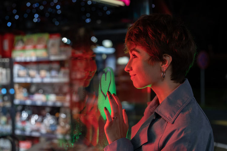 Cheerful attractive woman looks at shop window and draws heart on misted glass as sign of love