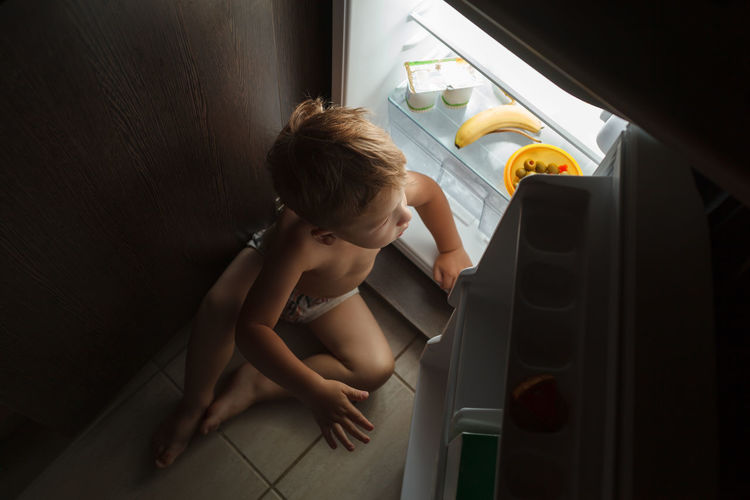 Cute boy sitting by refrigerator in house at night