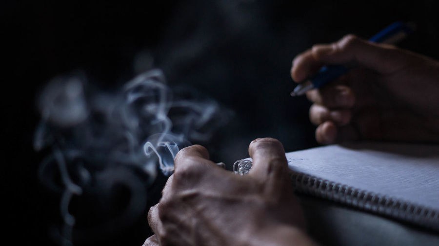 Cropped image of hand holding pen on book while smoking cigarette
