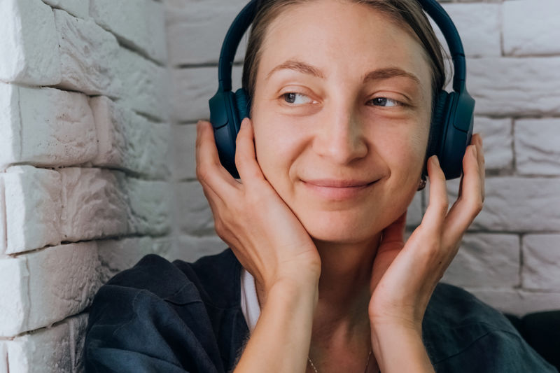 Smiling woman listening to music or podcast in wireless headphones while sitting near brick wall