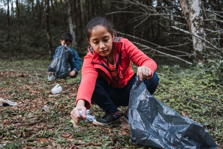 Ethnic volunteers with plastic bags picking rubbish from terrain against trees in summer woods in daylight