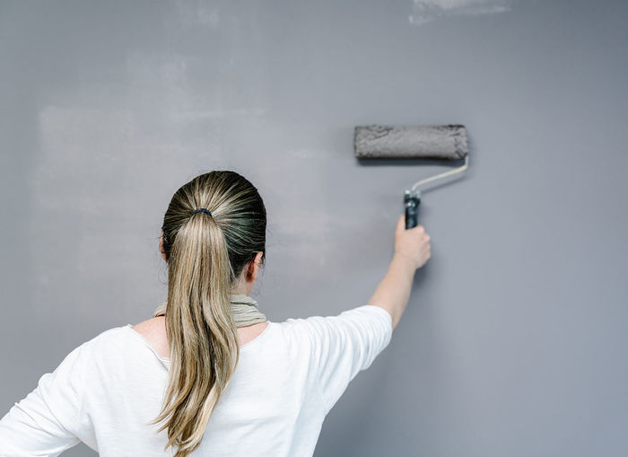 Backwards woman catching a painting roller full of grey painting on a wall. the painter is upping and downing the roller covering the wall with grey painting what remains wet. horizontal pic