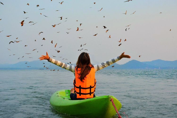 Rear view of woman with arms outstretched while kayaking on sea against birds in sky