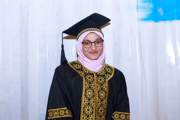 Portrait of smiling young woman wearing mortar board while standing against wall