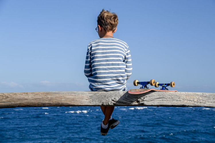 Rear view of boy sitting on wooden log over sea against clear blue sky