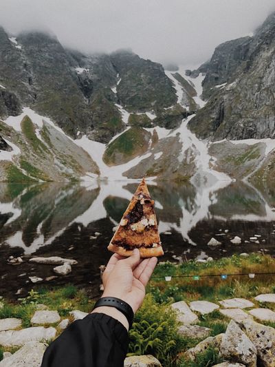 Cropped hand of person holding pizza slice against mountains during winter