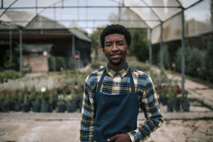 Smiling young farm worker standing against greenhouse