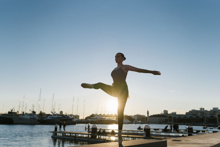 Young female dancer balancing on one leg while practicing at marina during sunset