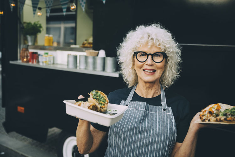 Portrait of senior owner with street food standing against commercial land vehicle in city