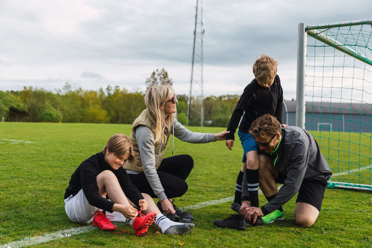 Parents helping teenage sons putting on football boots while preparing for playing soccer in green field