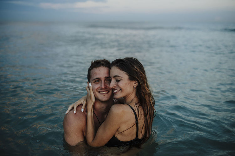 Smiling couple embracing while standing in water at beach during sunset