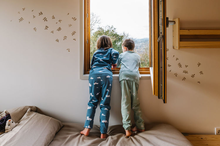 Full body back view of anonymous curios children in nightwear looking out window while standing on comfortable bed in bedroom