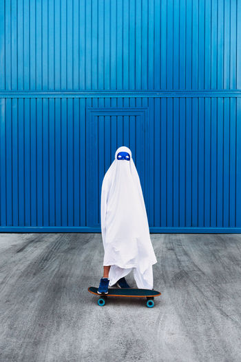 Unrecognizable child wearing mask and white ghost costume standing on skateboard in city during halloween and looking at camera