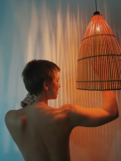 Rear view sensuous shirtless woman standing by lighting equipment against wall