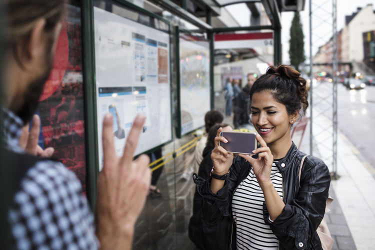 Smiling woman photographing man through smart phone at bus stop in city