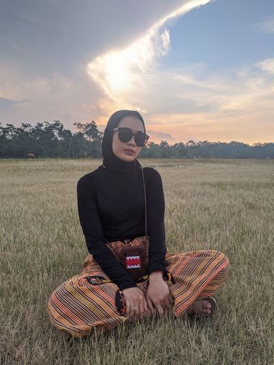 Woman wearing sunglasses sitting on land against sky during sunset