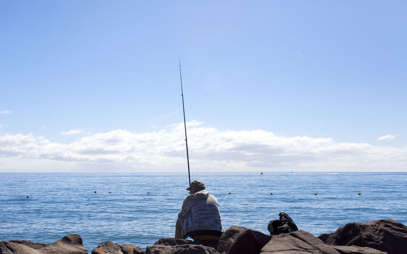 Rear view of man fishing while sitting on rocks by sea against sky