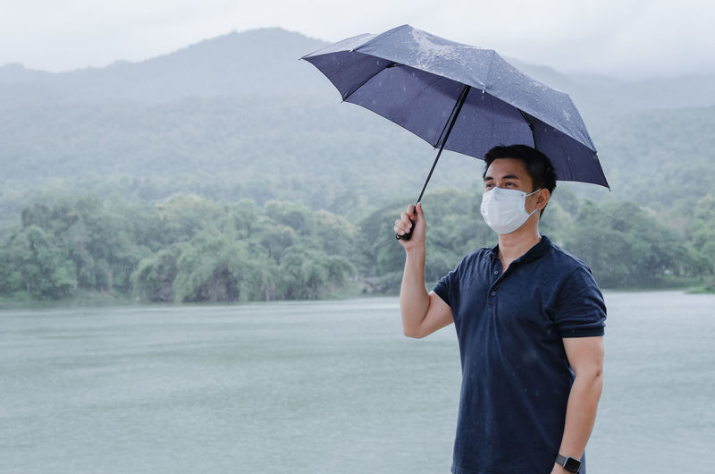 Asian man wearing face mask to protect from virus holding umbrella when raining in monsoon season.