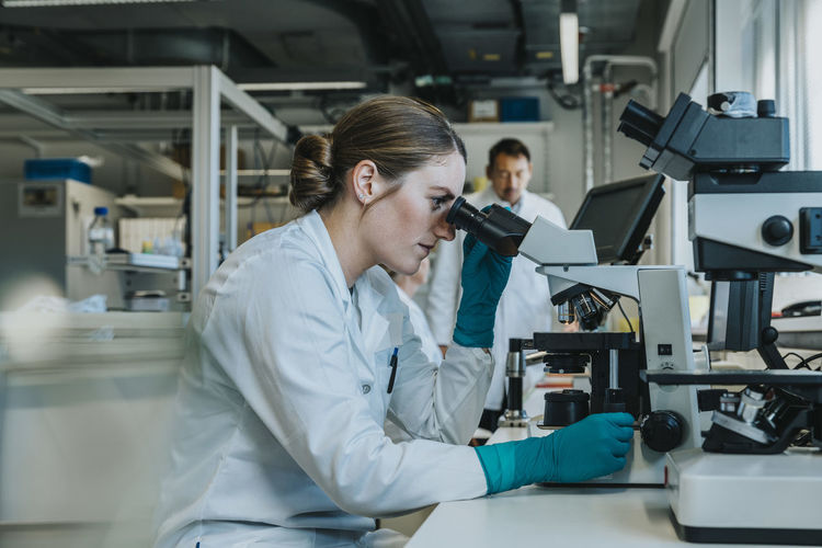 Young woman analyzing human brain microscope slide under microscope while sitting with scientists in background at laboratory