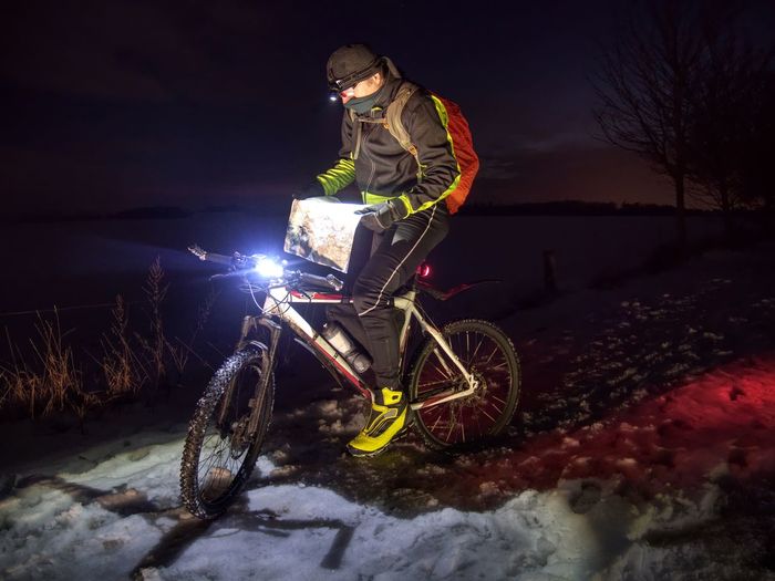 Young cyclist bow head in evening checks map in hands during winter night. snowy ground in darkness