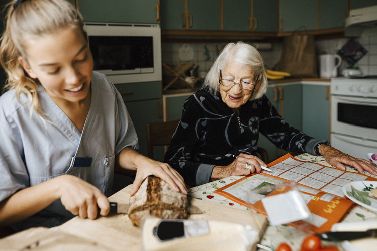 Young female healthcare worker cutting bread loaf sitting by senior woman solving sudoku at home