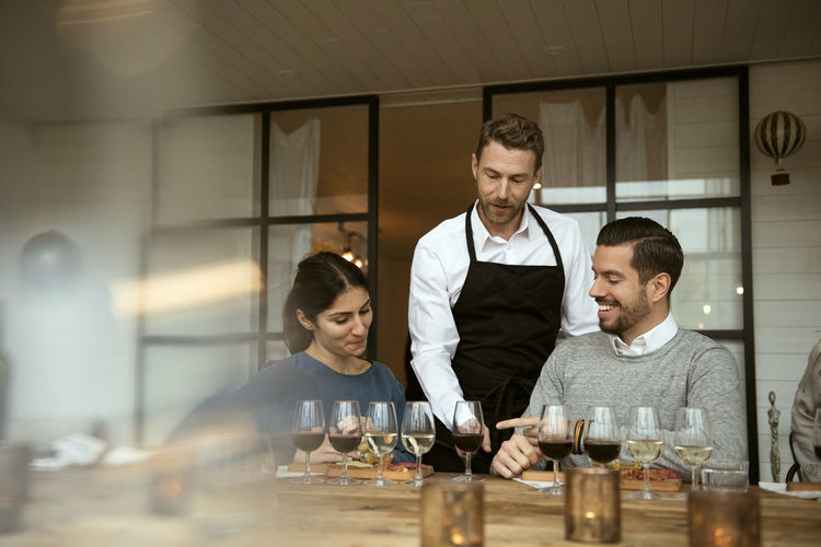 Man wearing apron standing by business people while pointing at wineglasses on table
